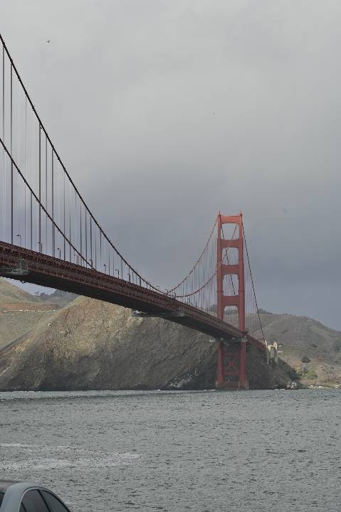 Red Golden Gate Bridge tower and suspension cables span over gray water toward green hills under a cloudy sky, with a car roof visible at the bottom edge.